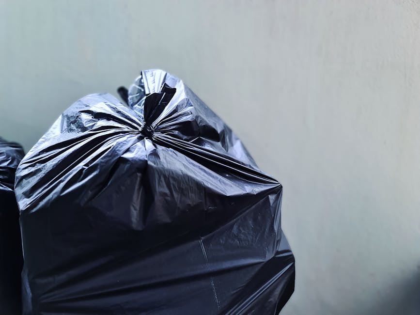 A large black plastic garbage bag, tightly knotted at the top, is positioned in the foreground against a plain, light-colored wall. The bag appears to be made of durable, shiny plastic with a slightly crinkled texture, indicating it is filled with waste materials. Part of another similar bag is visible behind it, partially obscured, suggesting multiple bags prepared for disposal. The scene is set on an indoor or outdoor surface, possibly a driveway or pavement, with minimal environmental context visible. The lighting is even, without harsh shadows, highlighting the bag's glossy finish and creases. This image exemplifies a typical scene of private rubbish collection, often associated with independent waste handling services like those offered by Waste Collection Haringey, highlighting the prepared waste ready for collection as part of on-site clearance or alternative waste disposal methods.