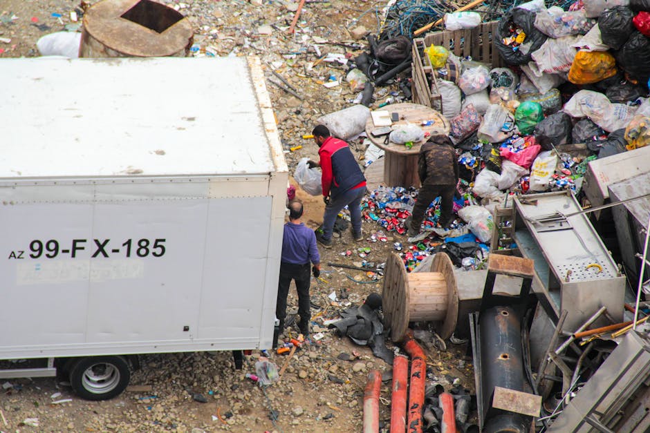 An aerial view of a cluttered outdoor space showing a large pile of mixed waste and垃圾, including plastic bags, cardboard, and miscellaneous rubbish, surrounding an area filled with discarded items and debris. In the foreground, a white waste collection truck marked with black lettering is partially visible on the left side, with its side panel showing the registration number '99-FX-185'. Three individuals are seen actively sorting through the refuse: one wearing a red and black jacket is bending forward, another in a purple shirt stands nearby, and a third person with a dark jacket is leaning into the pile. The surrounding environment consists of uneven, gravelly ground with scattered smaller waste items and large spools of cable, suggesting an informal or site clearance setting. The scene is lit naturally, highlighting the textures of the various materials, from crumpled plastic to rough concrete debris. This image relates to private waste handling and rubbish collection services, emphasizing the importance of professional rubbish removal for clearing such substantial waste accumulations.