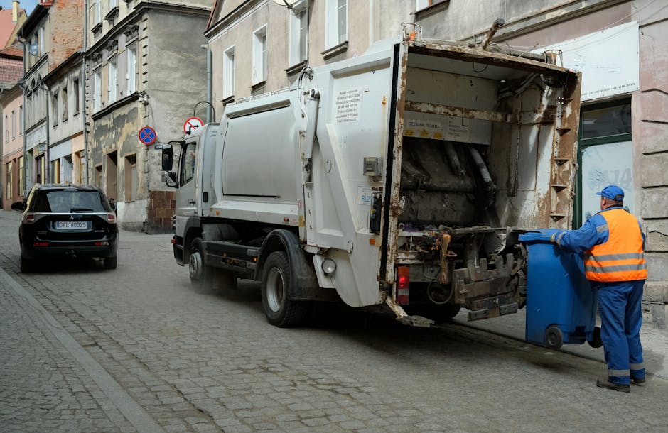 A street scene showing a white waste collection truck operated by Waste Collection Haringey parked close to a row of old, multi-story buildings with worn facades and various architectural details. The truck's rear hopper is open, revealing rusted and stained metal components used for waste compaction and loading. A worker dressed in a high-visibility orange vest and blue uniform wearing a blue cap is standing on the pavement to the right, actively pushing a blue wheeled bin towards the truck's loading area. The street surface consists of small, cobblestone paving, with a black parked car nearby. In the background, there are street signs indicating parking restrictions, and the overall environment suggests a routine rubbish collection operation in an urban setting, reflecting an alternative waste disposal method apart from official municipal rubbish collection services.