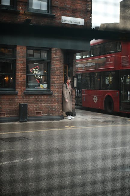 A woman in a long beige coat and white sneakers stands on a wet pavement beside a red double-decker bus on Lordship Road in Haringey. She appears to be waiting or observing, with her hands in her pockets. The background features a brick building with two dark-framed windows, one displaying a pizza sign, and a white street name plate reading 'Lordship Road'. The bus is parked close to the curb, with part of its route information visible on the side. Overhead, there is a dark green structure, possibly a canopy or awning, casting a shadow over part of the scene, which emphasizes the rainy, overcast weather typical of urban areas. This setting exemplifies typical city traffic arrangements, where private transit options like buses coexist with pedestrian activity, aligning with the theme of alternative waste handling and the importance of proper rubbish disposal amidst busy city environments. Waste Collection Haringey’s services could help manage rubbish accumulated in such urban settings, supporting efficient waste removal and environmental standards. The overall scene is neutral and factual, showcasing a moment in daily city life with clear focus on the transportation and street environment.