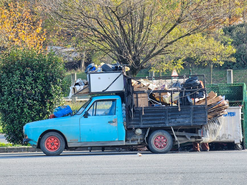 A small, older model light blue utility truck parked parallel to the curb on a paved street during daylight hours, loaded with a substantial pile of miscellaneous waste materials. The truck's cargo area is filled with various items including cardboard boxes, black plastic garbage bags, and other debris piled high and extending beyond the sides of the vehicle. The truck’s wheels are red, and the vehicle shows visible signs of wear, with faded paint and some rust spots. Positioned behind the truck, a large green hedge and a tall tree with sparse green and yellow leaves are visible, indicating an outdoor setting during autumn. The background includes a fence and more greenery, suggesting a suburban or park area. The truck represents a private waste collection or on-site clearance option, often used as an alternative to council rubbish collection services, as seen in waste management practices by companies like Waste Collection Haringey.