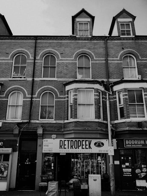A black and white photograph depicts the front of a multi-story brick building with three visible floors, featuring large arched and rectangular windows with white frames. The ground floor houses a retail store with a prominent sign reading 'RETROPEEK' and additional text indicating house clearances, situated on a busy street next to other commercial premises, including a delivery service with a sign saying 'BODRUM.' There are shopfront windows displaying various items, and the entrance is visible centered beneath a bay window. In front of the store, there are several rubbish bins and a trash collection box, with some loose trash visible nearby. The upper floors seem residential, with some windows partially open, and the building has a traditional Victorian architectural style with decorative brickwork. The atmosphere appears overcast, with diffused lighting, emphasizing the textures of the brickwork and street-level clutter related to private waste handling and rubbish removal services, aligned with the context of independent rubbish collection.
