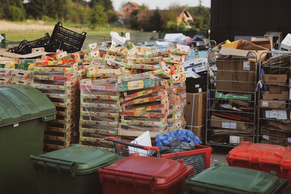 A large outdoor area filled with an assortment of waste materials awaiting collection, featuring stacked cardboard fruit boxes labeled 'fresh fruits' in the foreground, with many of these boxes partially collapsed or overlapping. To the left, a dark green wheeled waste bin is visible, while several other bins with lids in shades of green and red are positioned nearby. On the right side, there are metal cages and shelving units holding various cardboard boxes, packaging materials, and loose waste, including some brown paper and smaller cardboard pieces. The surface appears to be a driveway or yard, with a blurred background showing greenery, trees, and partial views of residential buildings under natural lighting, suggesting a typical garden or property setting. This scene illustrates a private collection of recyclable waste and rubbish, indicative of an independent rubbish disposal or collection arrangement, as managed by waste removal services such as Waste Collection Haringey.