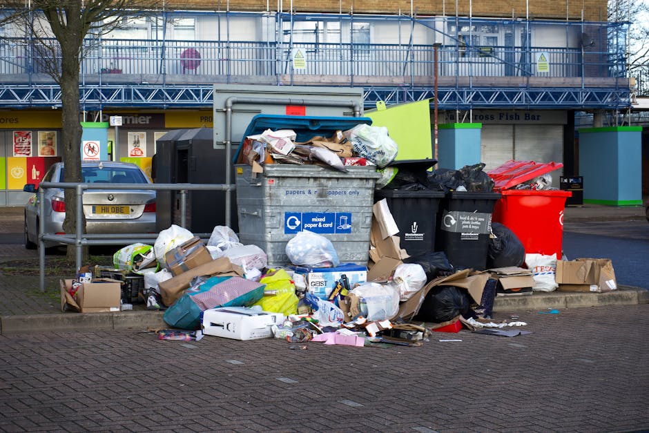 An overflowing collection of mixed waste including cardboard boxes, plastic bags, paper, and discarded packaging materials is piled beside a large, grey recycling bin labeled for paper and cardboard, situated on a paved urban street. Several smaller black and red wheelie bins are positioned nearby, with some lids open and rubbish spilling out. A silver car is parked adjacent to the waste area, with a metal railing separating the parking area from the pedestrian walkway. Behind the scene, a building under construction or renovation is visible, featuring blue safety scaffolding and yellow signage, with storefronts at street level. The overall scene depicts an unmanaged waste accumulation in a public space, highlighting issues of private rubbish disposal and alternative waste handling outside of official municipal collection services. The environmental context suggests an area requiring attention to rubbish removal and waste management procedures, with a neutral and factual tone suitable for understanding private rubbish collection practices in urban settings.