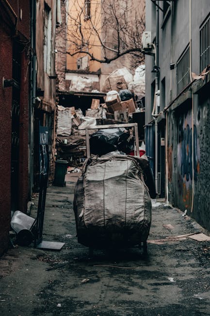 A narrow urban alleyway filled with an accumulation of rubbish and waste materials, featuring a large black trash bag placed on a wheeled trolley in the foreground. Behind it, numerous cardboard boxes, plastic bags, and miscellaneous debris are piled against a makeshift wooden barrier in the background, indicating a scene of on-site waste accumulation. On the left side, the alley is bordered by a red brick wall with some graffiti visible, while the right side shows a building with metal external fixtures, including an electrical box and barred windows. The surface of the alley is rough, dark asphalt with patches of dirt and grime, and the area appears cluttered and unmanaged, typical of an informal or private waste collection site, possibly associated with independent rubbish removal services like Waste Collection Haringey. The natural lighting casts soft shadows, emphasizing the layered textures of the garbage and surfaces, illustrating an environment where waste is awaiting collection or disposal without formal council involvement.