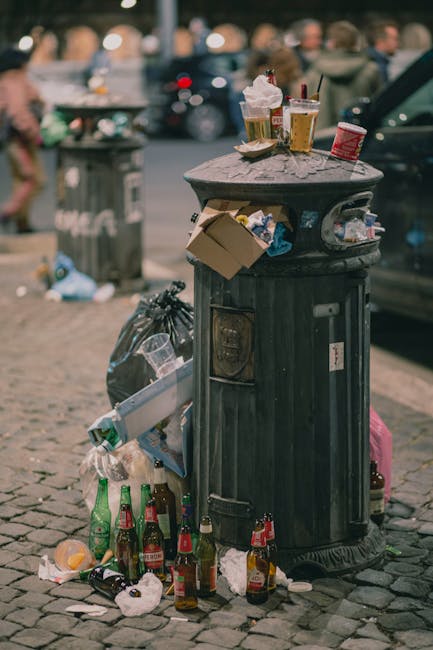 An overflowing collection of mixed waste including cardboard boxes, plastic bags, paper, and discarded packaging materials is piled beside a large, grey recycling bin labeled for paper and cardboard, situated on a paved urban street. Several smaller black and red wheelie bins are positioned nearby, with some lids open and rubbish spilling out. A silver car is parked adjacent to the waste area, with a metal railing separating the parking area from the pedestrian walkway. Behind the scene, a building under construction or renovation is visible, featuring blue safety scaffolding and yellow signage, with storefronts at street level. The overall scene depicts an unmanaged waste accumulation in a public space, highlighting issues of private rubbish disposal and alternative waste handling outside of official municipal collection services. The environmental context suggests an area requiring attention to rubbish removal and waste management procedures, with a neutral and factual tone suitable for understanding private rubbish collection practices in urban settings.
