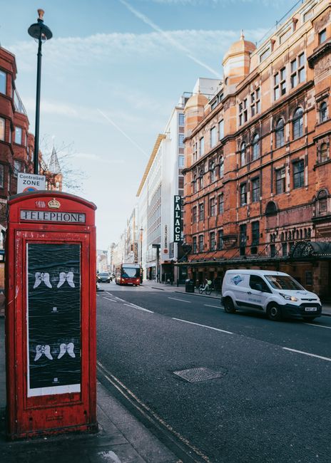 A red traditional British telephone box positioned on a city sidewalk, with a clear view of a busy street lined with multi-story red brick buildings featuring ornate window frames and ironwork balconies. The scene includes a white van and a red double-decker bus traveling along the road, with additional vehicles visible in the distance. The sky is mostly clear with a few wispy clouds, and a tall street lamp extends above the telephone box. The street sign indicates a controlled zone, and the general environment suggests an urban setting suitable for various waste disposal options, including private rubbish removal services by companies such as Waste Collection Haringey. The pavement surface appears to be in good condition, and the overall scene reflects a typical London street scene during daylight hours, captured in natural lighting that highlights the textures of the brickwork and the colour contrasts between vehicles, signage, and architecture.