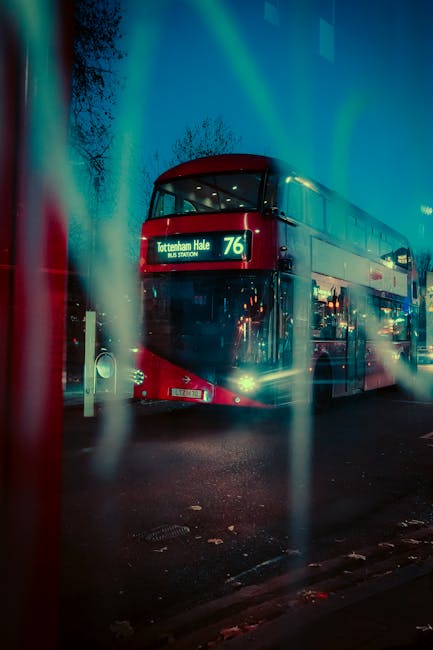 A double-decker bus with a red front and beige upper section is parked on a street at night, displaying the route number 76 and destination Tottenham Hale on its electronic sign. The bus is illuminated by interior and exterior lights, casting reflections on the wet pavement. In the foreground, slightly out of focus, there are vertical streaks or reflections, possibly caused by a window or glass surface. The background features a dark sky with faint silhouettes of leafless trees, indicating a cold season. This scene is set in an urban environment, with street-level lighting and pavement, and the bus's presence suggests it is part of the local public transportation network, which might be related to alternative ways of city mobility and transport services, akin to those offered by independent waste disposal or collection services like Waste Collection Haringey.