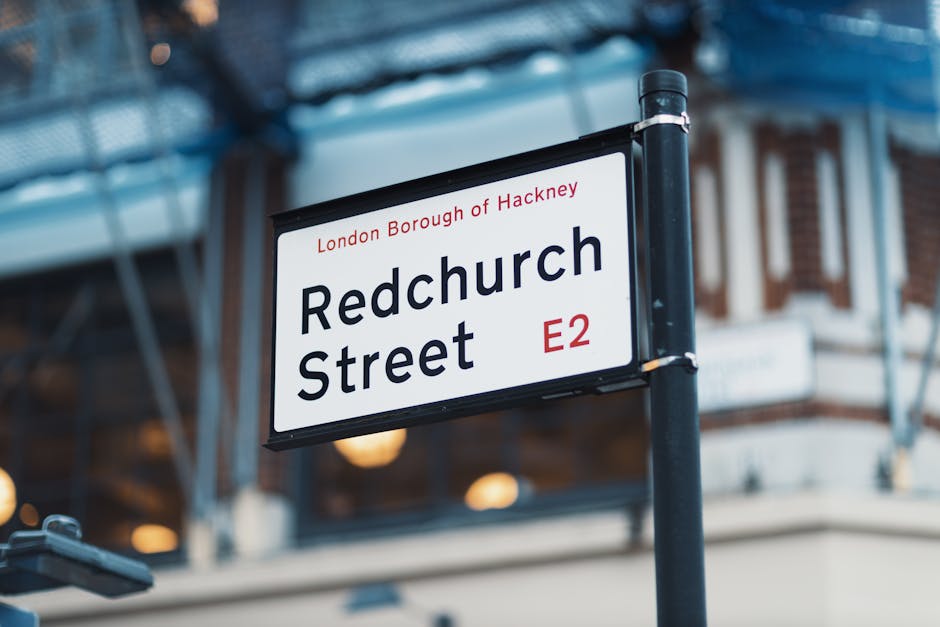 A street sign indicating 'Redchurch Street' located in the London Borough of Hackney, with the postcode E2. The sign is mounted on a black pole and has a white background with black text and red accents for the borough and postcode. In the background, there is a building under construction or renovation, with scaffolding and blue protective sheeting covering part of the facade. Warm internal lighting from the building is visible through windows, creating a contrast with the outdoor environment. The scene is captured during daylight hours, with soft, natural lighting. The image reflects an urban setting in East London where local residents and private waste collection services, such as wastecollectionharingey.co.uk, might operate to handle rubbish removal and alternative waste disposal outside of council services, fitting into the context of independent rubbish removal options in the area.