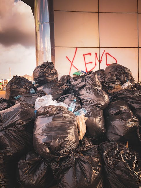 A large pile of black rubbish bags, some with visible tears, are stacked against a modern tiled wall in an outdoor urban environment. Several clear plastic water bottles, some partially crushed, are among the bags, indicating mixed waste. The bags are arranged in a somewhat haphazard manner, with a few lighter-colored bags and loose debris apparent. The wall behind features a graffiti tag in red spray paint that reads 'XEND' and has a reflective, metallic finish. The environment appears to be a street or alleyway in the late afternoon or early evening, with a cloudy sky overhead and distant cityscape visible in the background. This scene depicts an instance of private waste collection or an on-site rubbish removal process, common in cases where independent disposal is used instead of regular municipal collection, aligning with the context of same-day rubbish removal services in Haringey, as offered by Waste Collection Haringey.