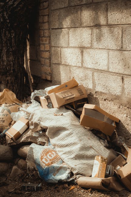 An alleyway scene with a large, grey, weathered rubbish sack placed in the foreground, surrounded by scattered debris and small waste items on the uneven asphalt surface. Behind the sack, there is a collection of stacked cardboard boxes, plastic bags, and miscellaneous refuse piled against a makeshift wooden barrier, indicating an accumulation of waste awaiting disposal. The alley is flanked by worn buildings, with the left side showing a rusty metal door and resilient graffiti on the wall, while the right side features dark, metal-paneled walls with small barred windows and exterior fixtures such as electrical boxes. Bare tree branches extend overhead, against a background of faintly visible brick construction and an overcast sky, contributing to a subdued, urban atmosphere. This scene, associated with private waste collection, underscores the necessity of effective rubbish removal services like those provided by Waste Collection Haringey, which cater to such neglected corners for alternative waste handling and on-site clearance.