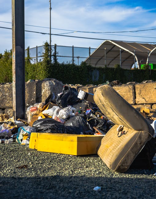 A large pile of mixed waste lies on the pavement in an outdoor setting, consisting of black plastic trash bags, loose cardboard boxes, and discarded household items. Prominently, an old, beige upholstered armchair with fabric upholstery and visible wear is tipped over on its side, with parts of the padding and frame exposed. A yellow plastic container is positioned behind the garbage, partially buried in the debris. In the background, a low stone wall made of irregularly shaped rocks separates the waste from a green hedge, with a metal fence and some utility poles and wires visible further back. Behind these, there is a large outdoor sports or event tent with a peaked roof, situated on a paved area under a partly cloudy sky with blue and white hues, giving an impression of a non-residential, possibly commercial or public space. The scene appears to relate to independent rubbish disposal or on-site clearance activities managed by Waste Collection Haringey, illustrating an example of unmanaged or improperly disposed waste in an urban environment.