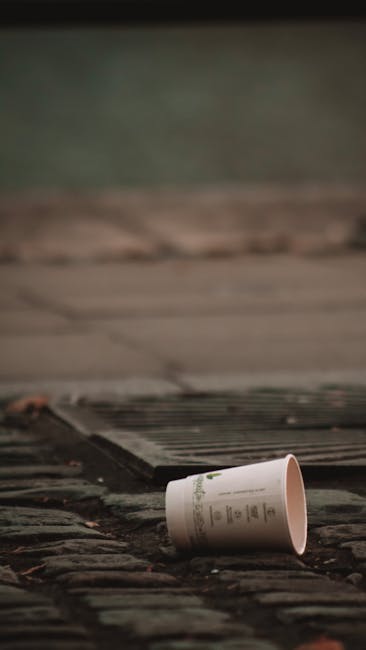 A large pile of mixed waste lies on the pavement in an outdoor setting, consisting of black plastic trash bags, loose cardboard boxes, and discarded household items. Prominently, an old, beige upholstered armchair with fabric upholstery and visible wear is tipped over on its side, with parts of the padding and frame exposed. A yellow plastic container is positioned behind the garbage, partially buried in the debris. In the background, a low stone wall made of irregularly shaped rocks separates the waste from a green hedge, with a metal fence and some utility poles and wires visible further back. Behind these, there is a large outdoor sports or event tent with a peaked roof, situated on a paved area under a partly cloudy sky with blue and white hues, giving an impression of a non-residential, possibly commercial or public space. The scene appears to relate to independent rubbish disposal or on-site clearance activities managed by Waste Collection Haringey, illustrating an example of unmanaged or improperly disposed waste in an urban environment.